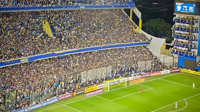 Fotbollsläktare fylld med folk, del av fotbollsplan, 3 spelare skymtas