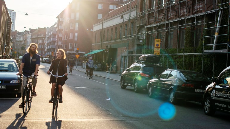 Två personer cycklar fram i Malmömiljö i skymning.